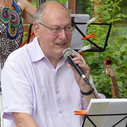 Ein älterer Mann sitzt draußen auf einem Stuhl und singt in ein Mirkofon. Vor ihm steht ein Notenständer. 