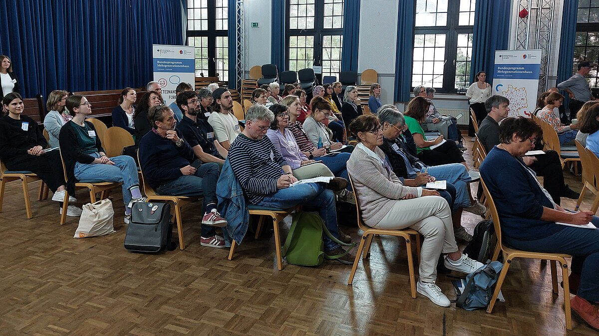 Mehrere Menschen sitzen in Stuhlreihen in einem großen Raum und folgen eine Rede