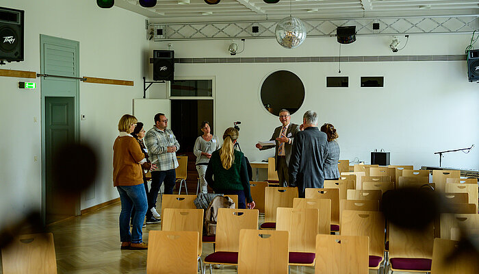 Eine Menschengruppe steht in einem Saal mit vielen Stühlen und unterhält sich