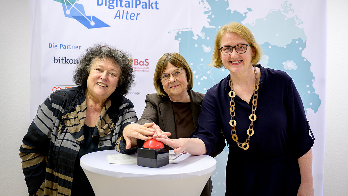 Drei Frauen stehen an einem Tisch, lächeln und drücken einen roten Buzzerknopf.