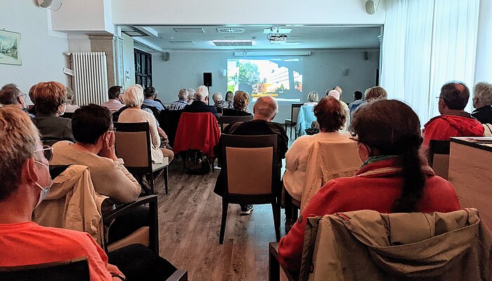 Eine große Gruppe Menschen sitzt auf Stühlen und schaut auf eine Leinwand