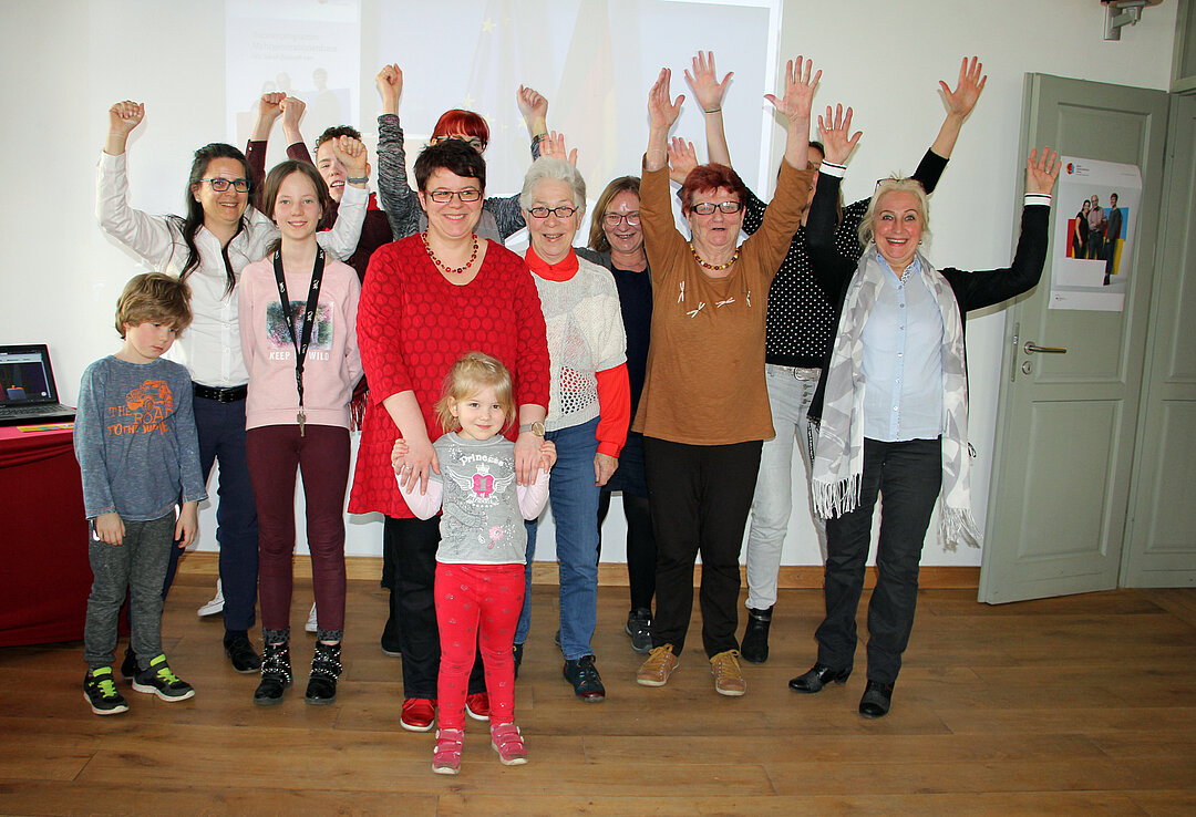 Auf dem Foto sind 12 PErsonen unterschiedlichen alters abgebildet, die die Arme in die Luft strecken.