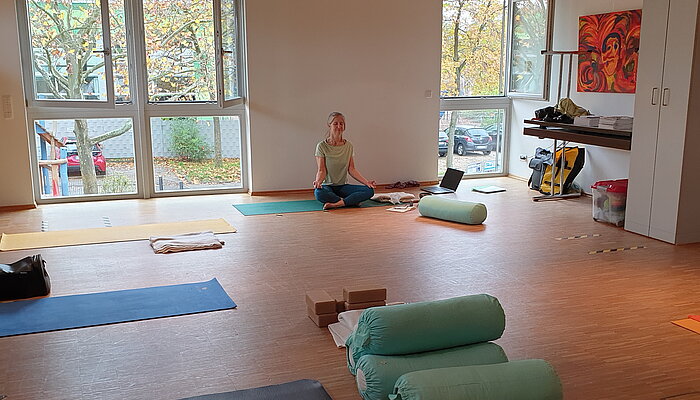 Eine Yogalehrerin sitzt in einem großen hellen Raum auf einem Meditationskissen.