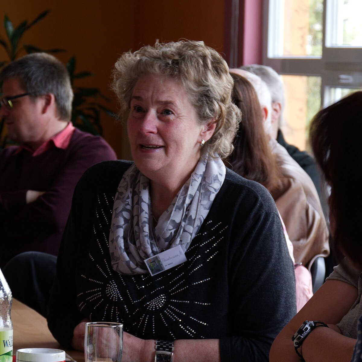Im Mittelpunkt des Bild sitzt eine mittelalte Frau an einem Tisch, sie ist umgeben von mehreren anderen Personen.