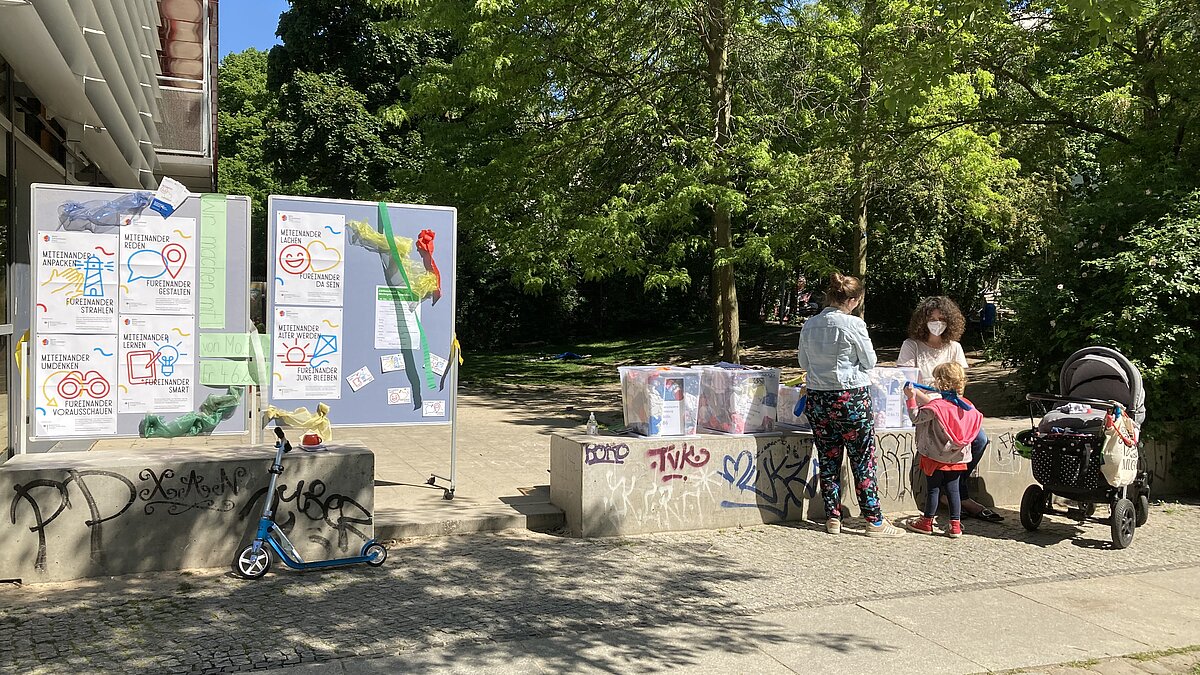 Zwei Metaplanwände und Kisten mit Kleidern stehen in einem Park, zwei Frauen und ein Kind unterhalten sich