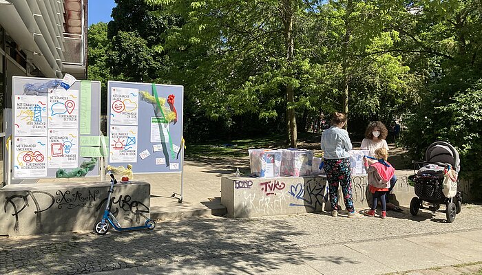 Zwei Metaplanwände und Kisten mit Kleidern stehen in einem Park, zwei Frauen und ein Kind unterhalten sich