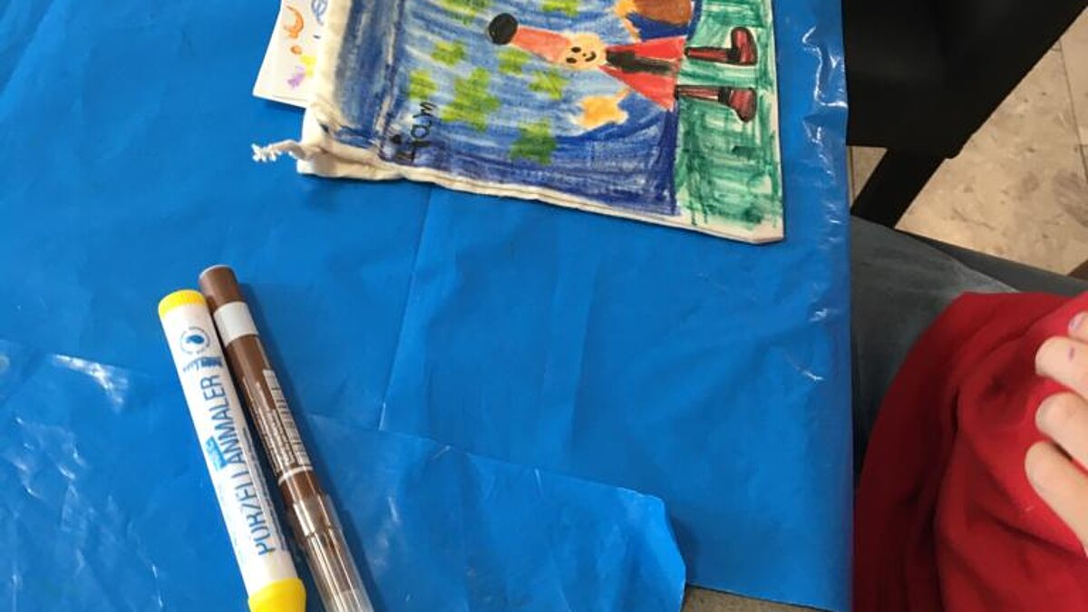 Eine Kinderhand greift nach einem Stift neben einer bunt bemalten Stofftasche.