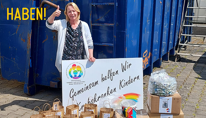 Eine Frau steht mit einem Pappschild mit der Aufschrift "Gemeinsam helfen wir krebskranken Kindern" hinter mehreren Papiertüten und zeigt den Daumen hoch.