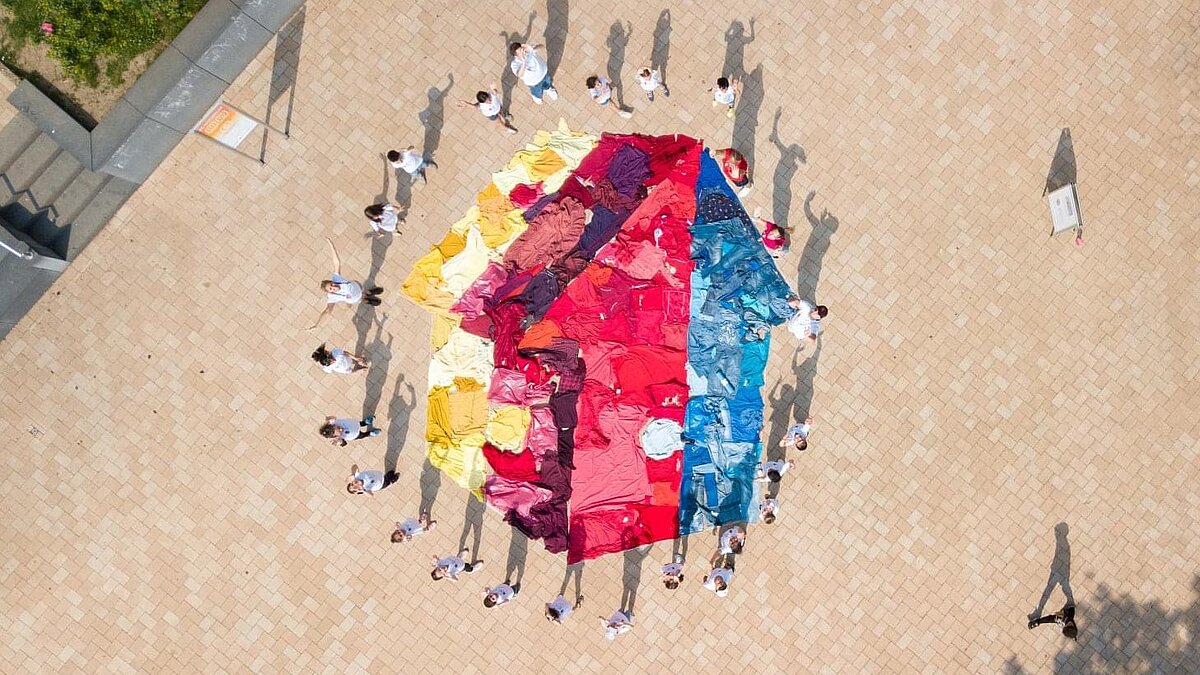 Aufnahme von oben: Viele Menschen in weißen T-Shirts stehen um ein aus gefärbtem Stoff auf den Boden gelegtes Mehrgenerationenhaus-Logo