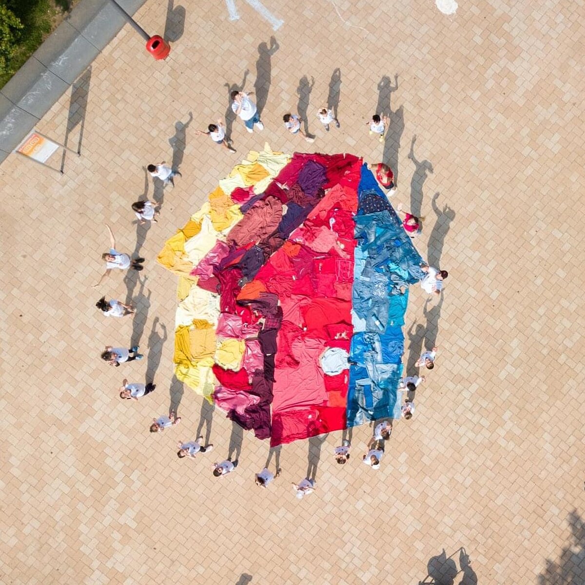 Aufnahme von oben: Viele Menschen in weißen T-Shirts stehen um ein aus gefärbtem Stoff auf den Boden gelegtes Mehrgenerationenhaus-Logo