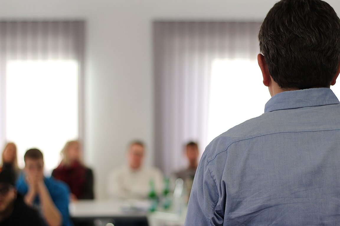 Person von hinten, die vor mehreren an Tischen sitzenden Menschen steht