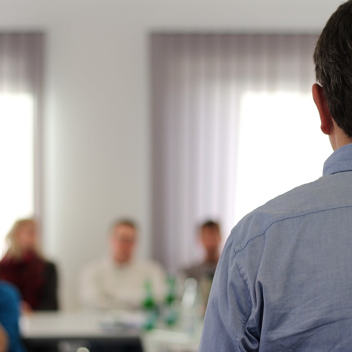 Person von hinten, die vor mehreren an Tischen sitzenden Menschen steht