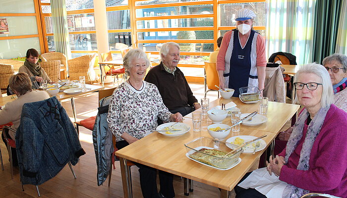 Drei ältere Frauen und ein Mann sitzen an einem Tisch mit Tellern, dahinter steht die Köchin