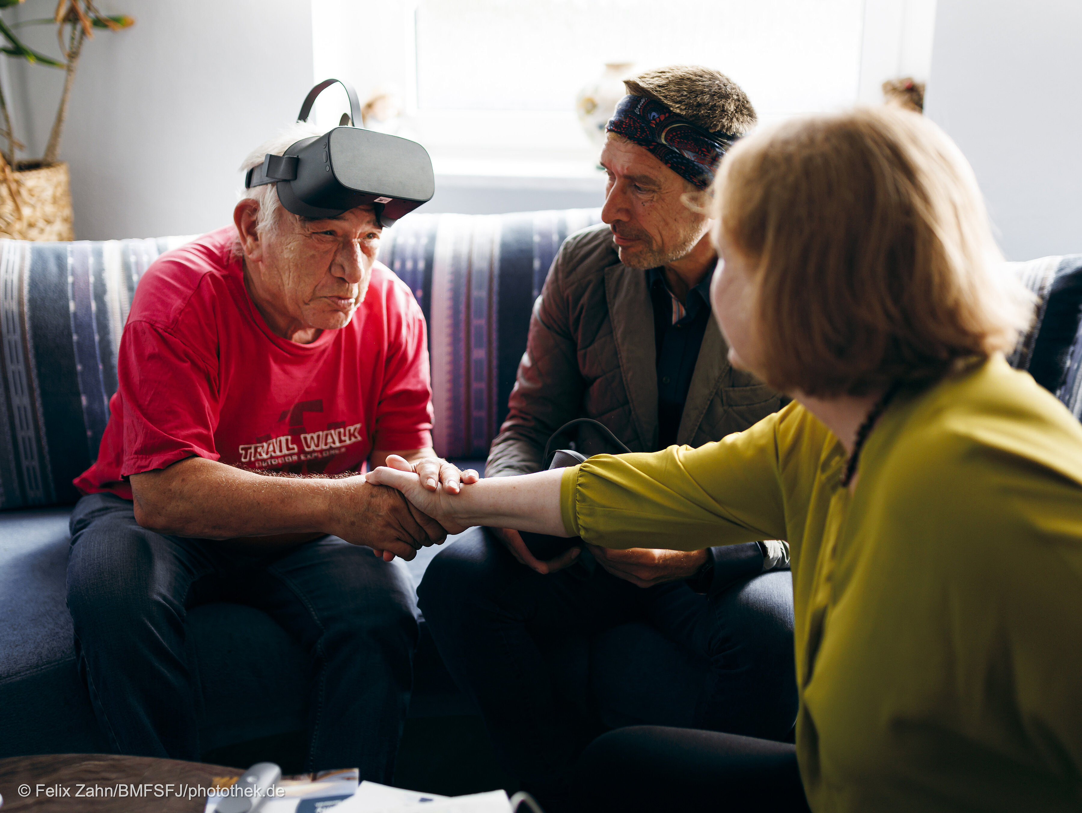 Bundesfamilienministerin Lisa Paus reicht einem älteren Mann, der eine VR-Brille auf der Stirn trägt, die Hand, ein weiterer älterer Herr sitzt daneben.