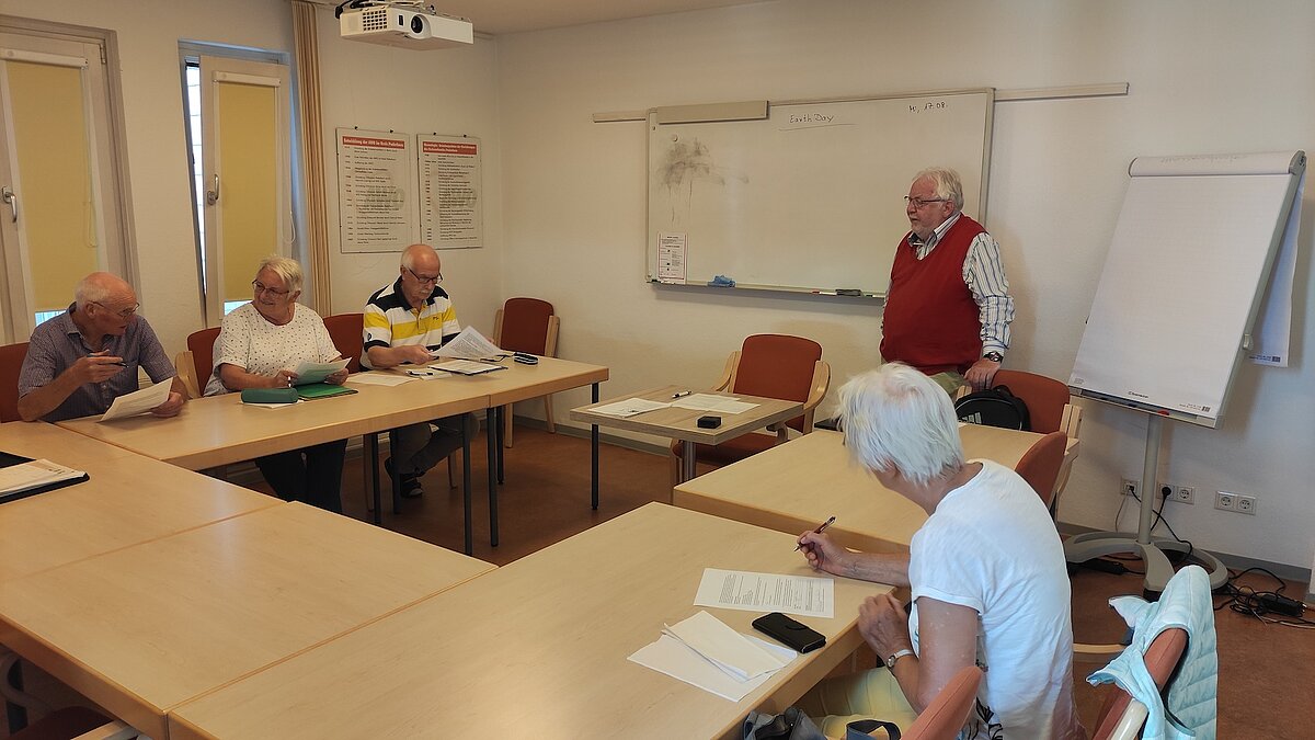 Zwei ältere Herren und zwei ältere Damen sitzen an Tischen und füllen Arbeitsblätter aus. Ein älterer Herr steht an der Tafel.