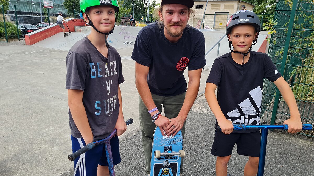Drei Jungs tragen Helme, stehen auf ihren Rollern und lächeln.