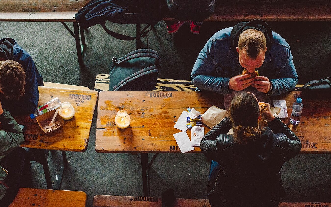 Drei Menschen sitzen an Biertischen und essen, Perspektive von oben auf die Köpfe