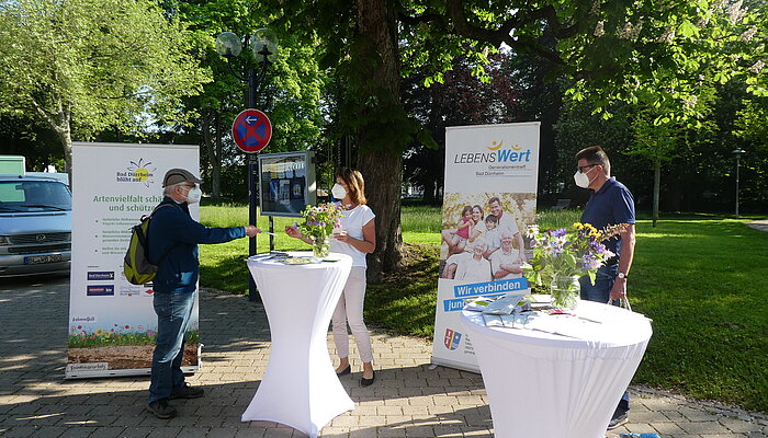 Drei Menschen stehen an zwei Stehtischen mit Informationsmaterialien und unterhalten sich