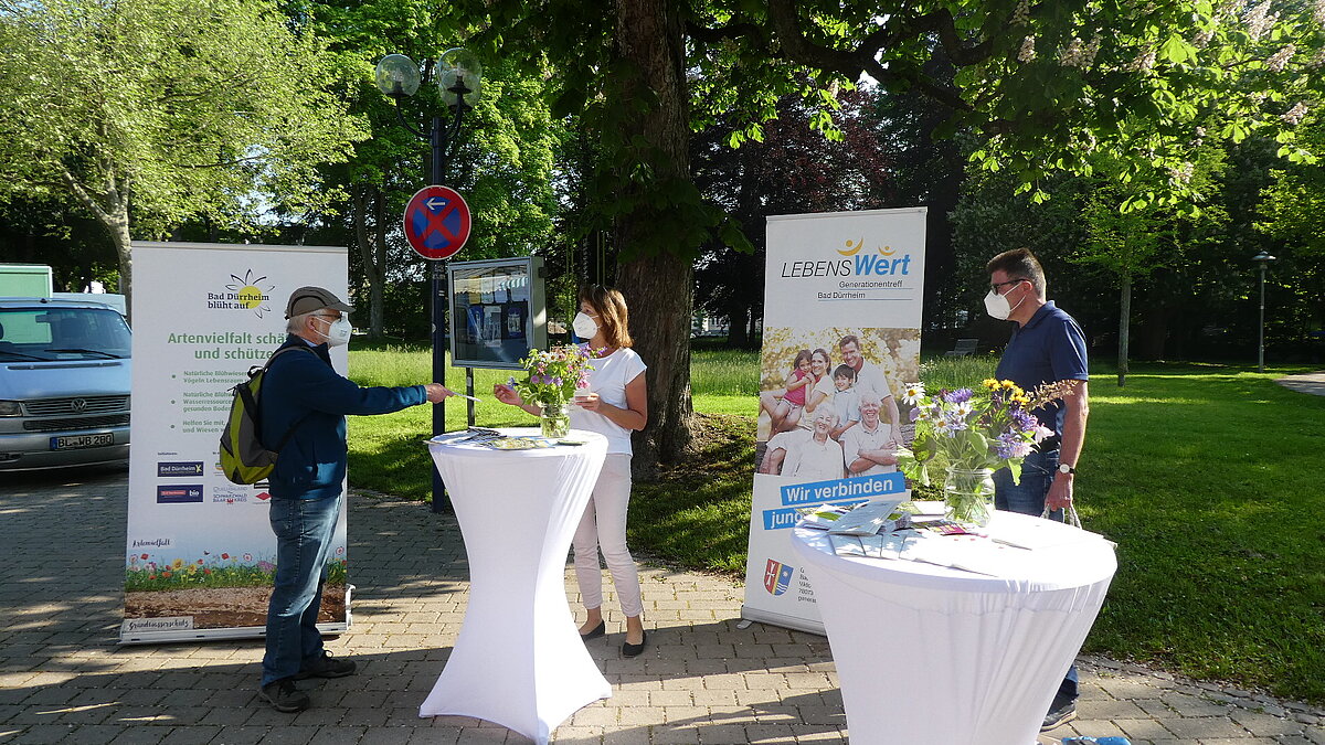 Drei Menschen stehen an zwei Stehtischen mit Informationsmaterialien und unterhalten sich