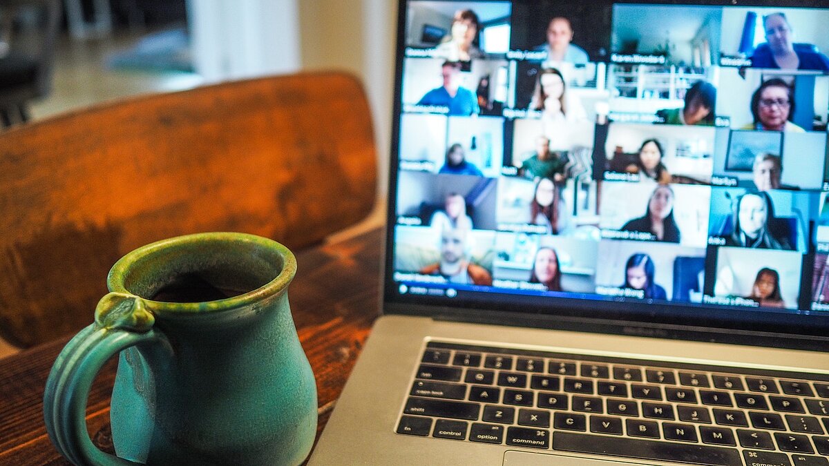 Laptop zeigt verschiedene Menschen in einer Videokonferenz, daneben steht eine Tasse