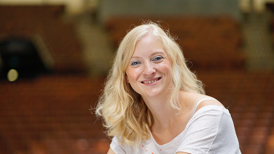 Eine blonde Frau sitzt in einem Saal und lächelt in die Kamera. Es ist Fenja Grieshammer, die sich ehrenamtlich im Mehrgenerationenhaus Wunsiedel engagiert. 