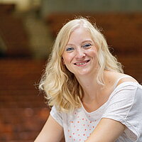 Eine blonde Frau sitzt in einem Saal und lächelt in die Kamera. Es ist Fenja Grieshammer, die sich ehrenamtlich im Mehrgenerationenhaus Wunsiedel engagiert. 