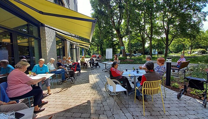 Vierzehn Menschen sitzen an Café-Tischen vor einem Gebäude, trinken Kaffee und unterhalten sich