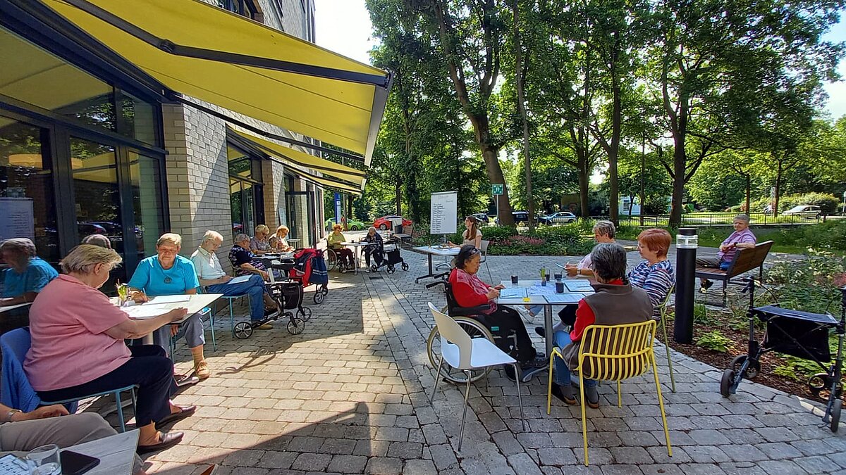 Vierzehn Menschen sitzen an Café-Tischen vor einem Gebäude, trinken Kaffee und unterhalten sich