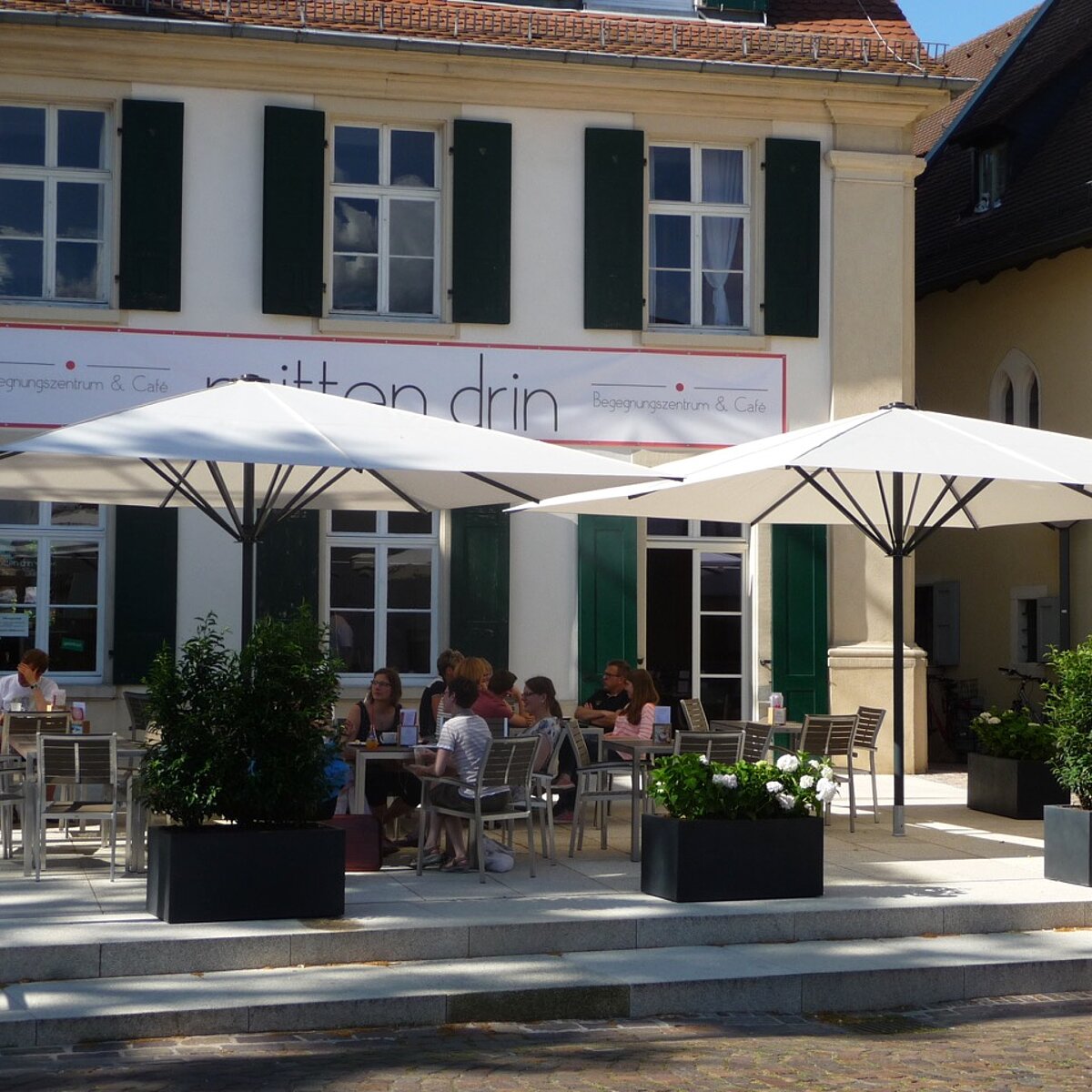 Vor dem Mehrgenererationenhaus gibt es eine großzügige Terrasse mit Stühlen und Tischen an den Menschen unter großen Sonnenschirmen sitzen.