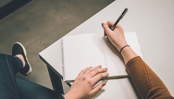 Hand hält einen Stift und schreibt in ein Notizbuch auf einem Tisch