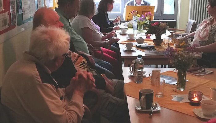 Sechs Menschen sitzen an einem Tisch und trinken Kaffe, am Kopfende spielt ein Mann auf einem Keyboard.