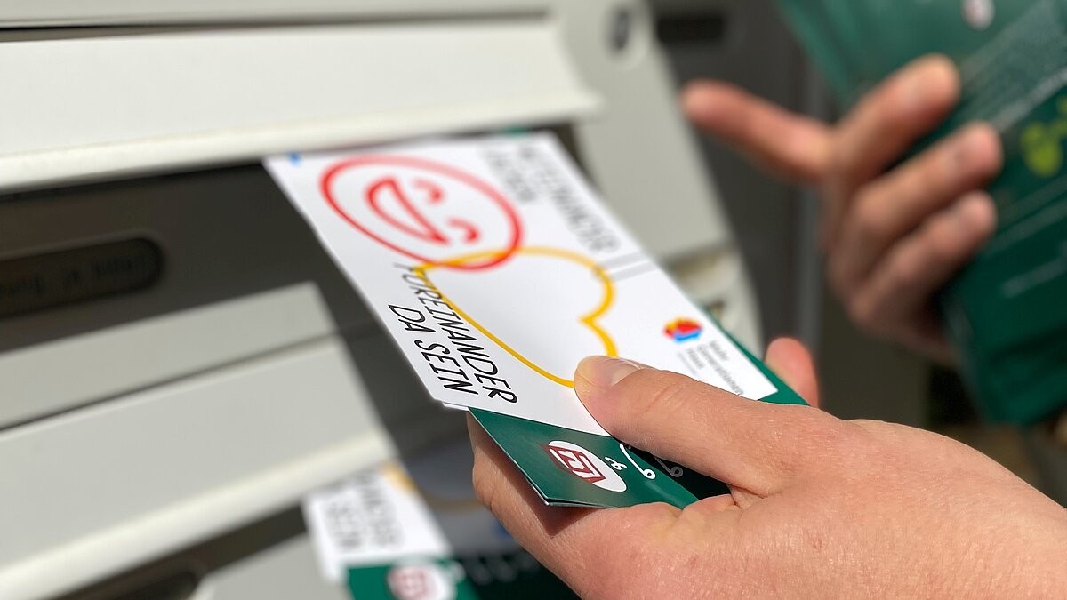Eine Hand steckt eine Postkarte in einen Briefkasten.