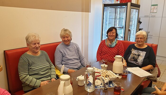 Vier ältere Damen sitzen gemeinsam an einem Tisch, auf denen Kaffeetassen und Kuchen stehen.