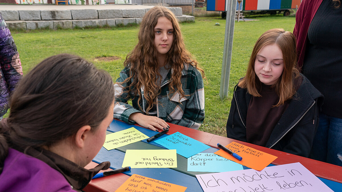 Drei Jugendliche sitzen an einem Tisch auf einer Wiese, auf dem Zettel mit verschiedenen Wünschen für Mädchen und Frauen auf der ganzen Welt