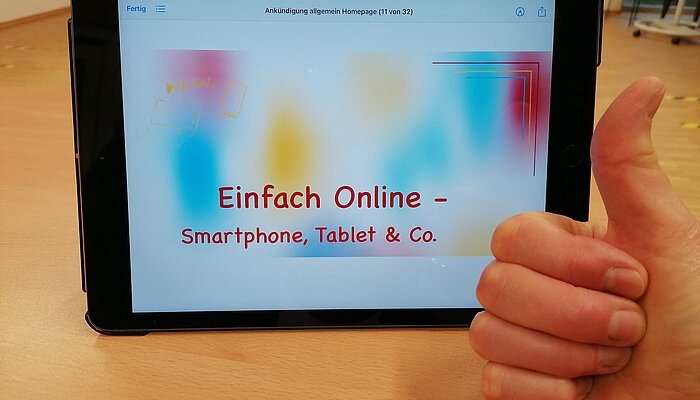 Ein Ipad steht aufrecht auf einem Tisch. Eine Hand zeigt "Daumen hoch". 