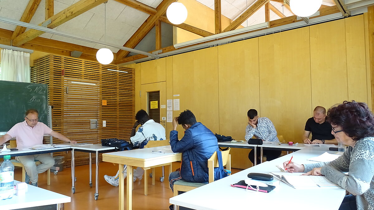 Eine Frau und vier Männer sitzen an Pulten, vorne sitzt der Lehrer an einem Pult