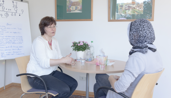 Zwei Frauen sitzen an einem Tisch und reden. Im Hintergrund steht eine weiße Tafel mit Notizen.