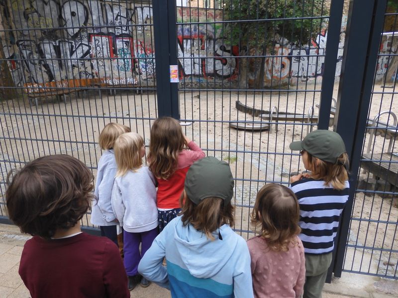 Sieben Kinder stehen vor dem Zaun eines Spielplatzes