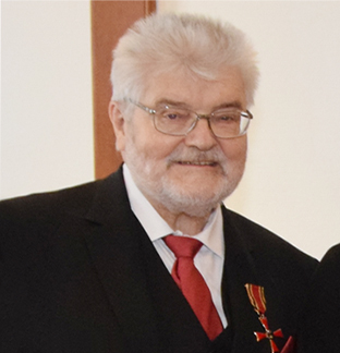 Das Foto zeigt einen älteren Mann mit grauen Haaren und grauem Bart sowie einer Brille. Er trägt einen schwarzen Anzug, ein weißes Hemd und eine rote Krawatte.