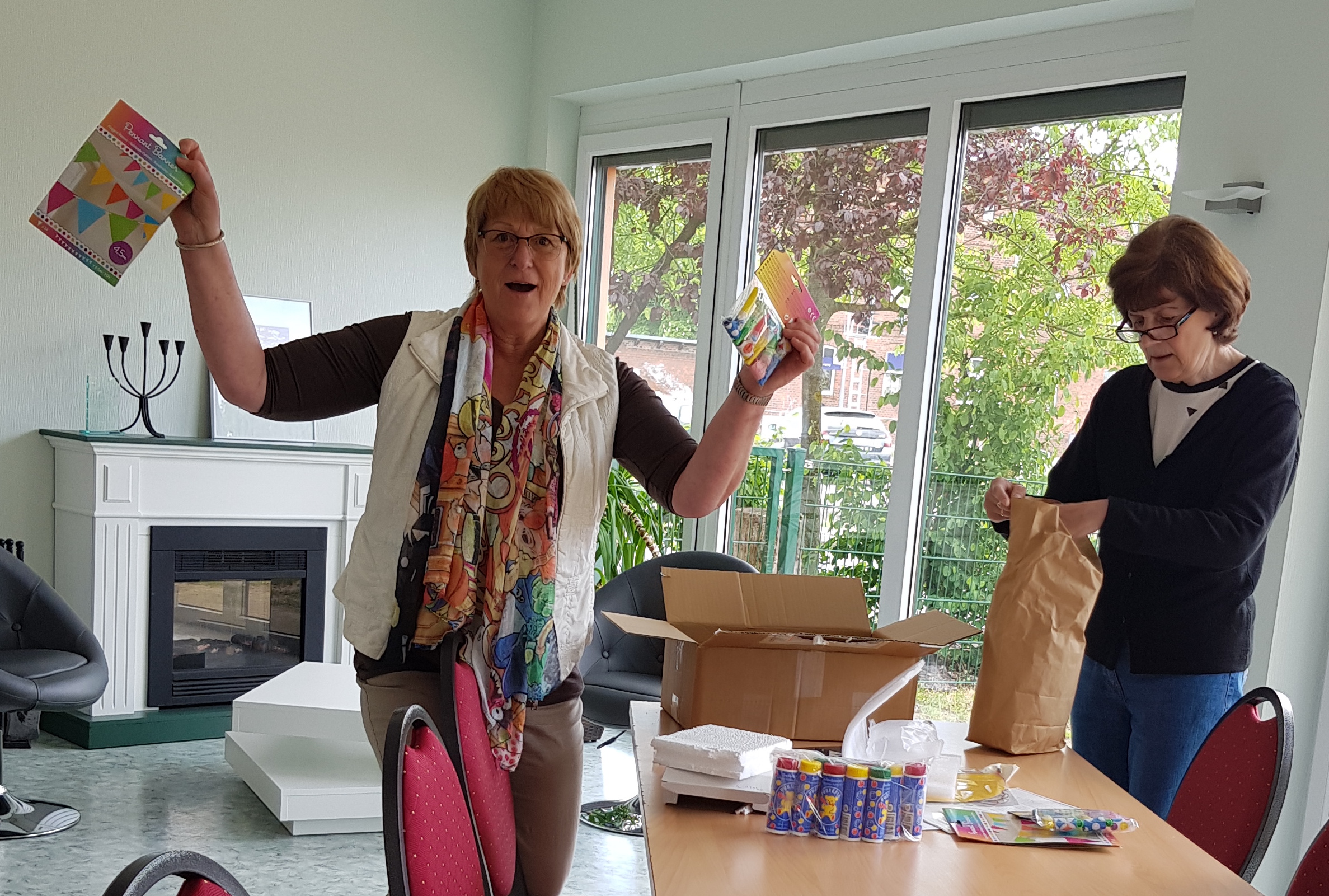 Zwei Frauen packen das Gewinnerpaket aus, die Frau links hält ein Paket mit Luftballons und eine Packung mit einer Girlande in den Händen und jubelt.